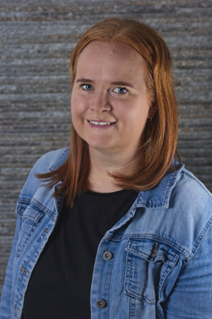 Bewerbungsfotos einer Frau mit schulterlangen, roten Haaren, blauer Jeansweste vor einem steinigen Hintergrund.