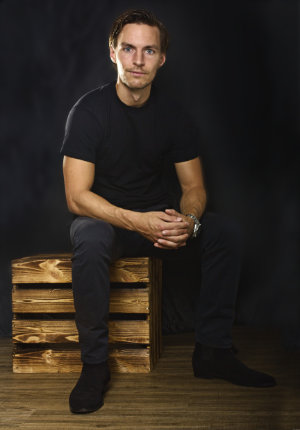 Ganzkörperportrait eines sitzenden Mannes auf einer Holzkiste vor einem schwarzen Hintergrund.