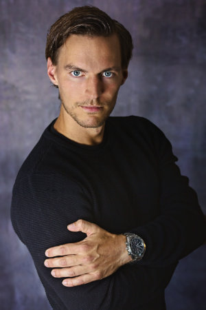 Bewerbungsfotos eines Mannes mit verschränkten Armen und graulich gemustertem Hintergrund.